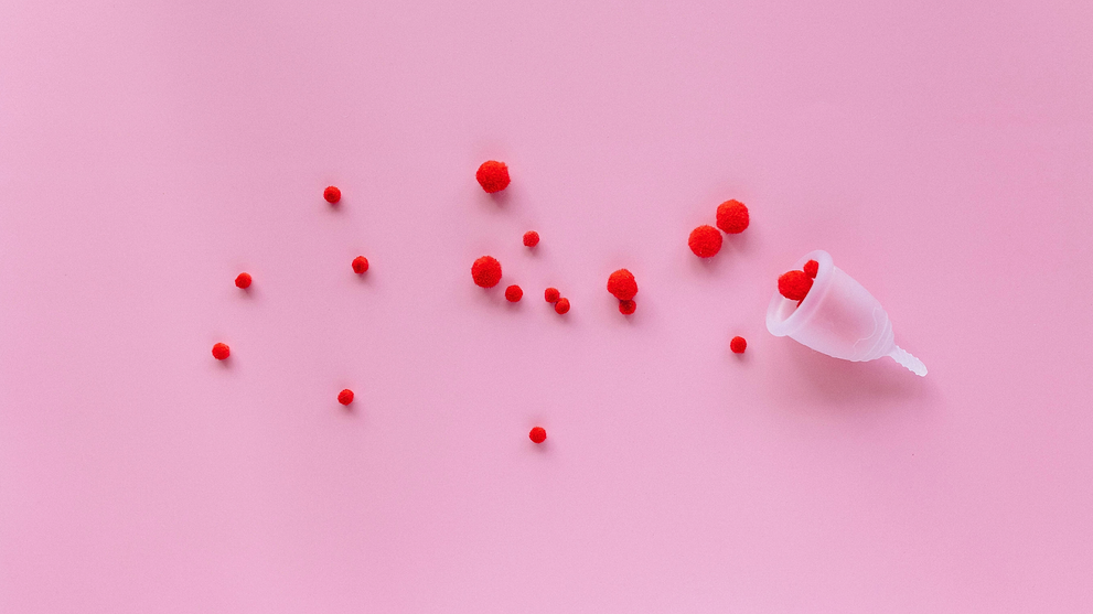 Menstrual cup tipped over on pink background with scattered red pom-poms symbolizing menstrual flow.