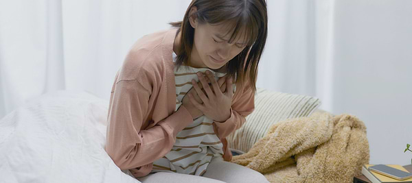 Woman sitting on a bed holding her chest, appearing distressed or experiencing chest discomfort.