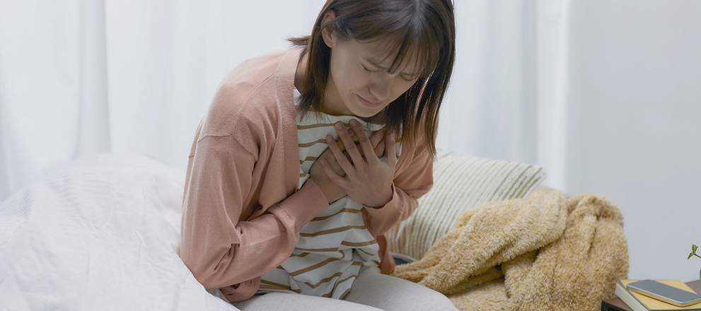 Woman sitting on a bed holding her chest, appearing distressed or experiencing chest discomfort.