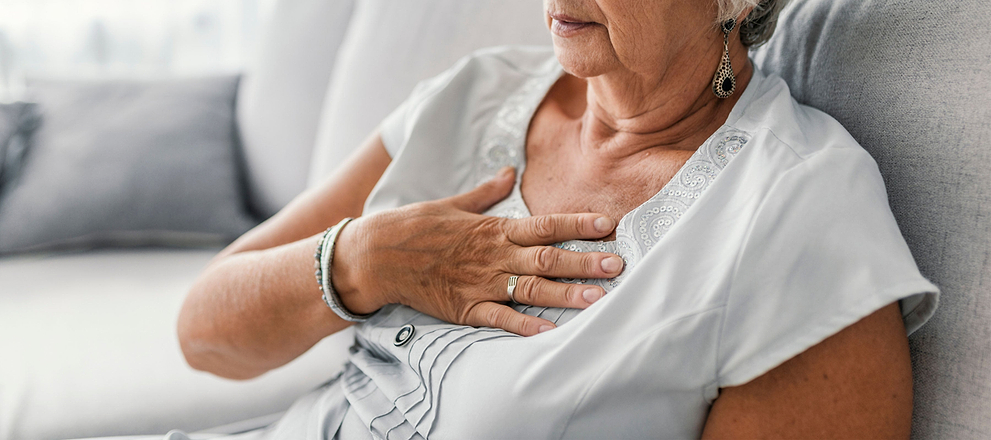 Older woman sitting on a couch with her hand on her chest, appearing uncomfortable or in pain.