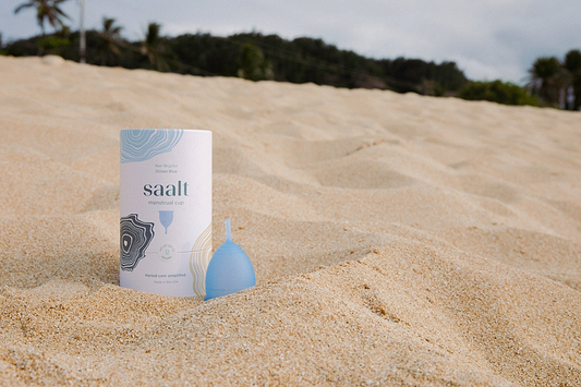 Saalt menstrual cup and packaging displayed on a sandy beach with palm trees in the background.