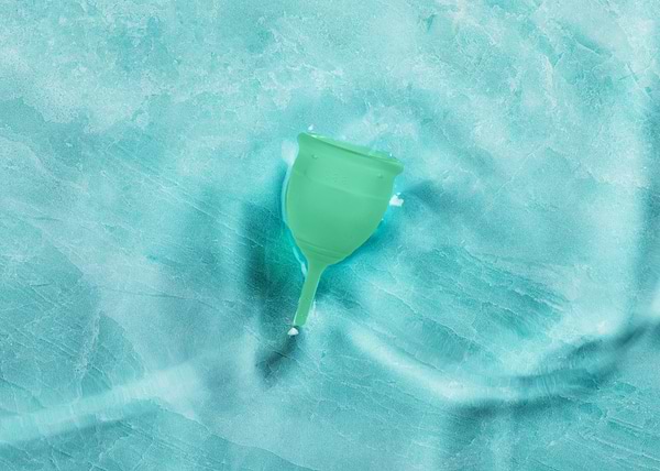 Green menstrual cup resting on a textured aqua surface with soft lighting and subtle shadows.