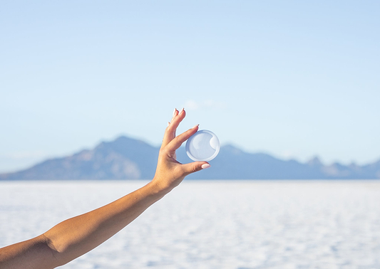 Hand holding a translucent menstrual disc against a bright sky and distant mountain landscape