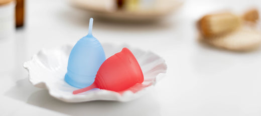 Blue and pink menstrual cups displayed on a white shell-shaped dish in a clean, minimalist setting.