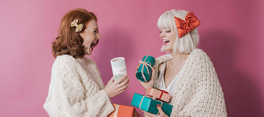 Two women exchanging gifts and holding Saalt products while smiling against a pink background.