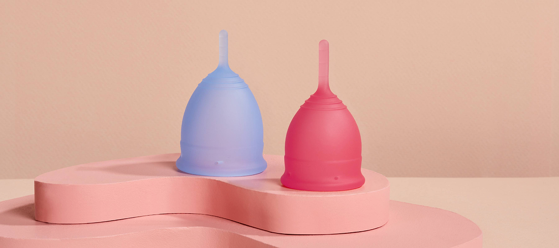 Blue and pink menstrual cups displayed on a soft pink pedestal against a neutral background.