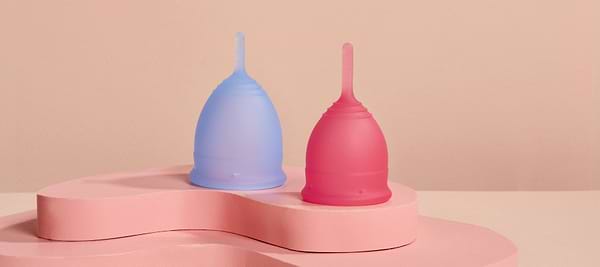 Blue and pink menstrual cups displayed on a soft pink pedestal against a neutral background.