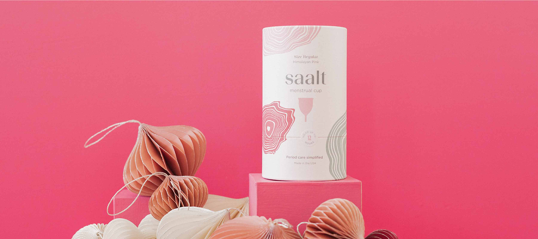 Saalt menstrual cup packaging displayed on pink blocks with decorative paper ornaments and a bright pink background.