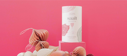 Saalt menstrual cup packaging displayed on pink blocks with decorative paper ornaments and a bright pink background.