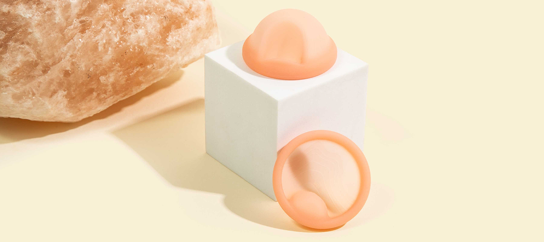 Peach-colored menstrual discs displayed on a white cube beside a textured salt rock in soft natural light.