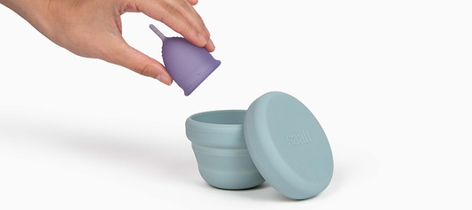 Hand placing a purple menstrual cup into a blue collapsible Saalt storage container on a white background.