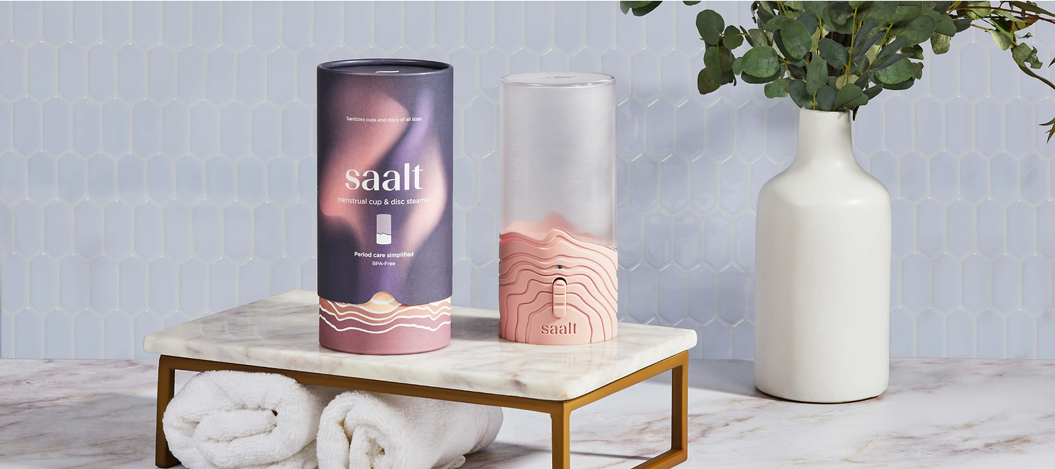 Saalt menstrual cup steamer and packaging displayed on a marble bathroom counter beside a white vase.
