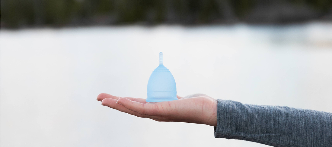 Hand holding a light blue menstrual cup outdoors against a soft blurred background.