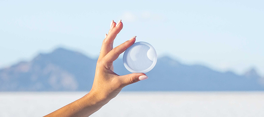 Hand holding a translucent menstrual disc against a soft blue mountain landscape background.