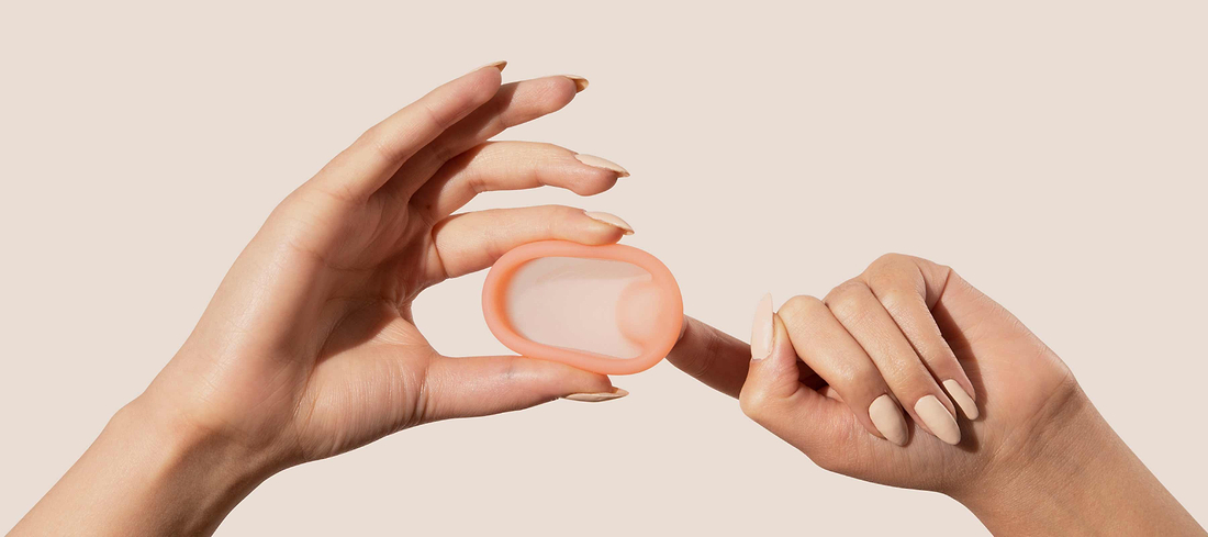 Hands holding and pointing to a peach-colored menstrual disc against a neutral background.