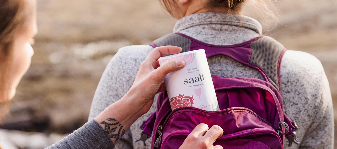 Person placing a menstrual cup box into a backpack during an outdoor hike.