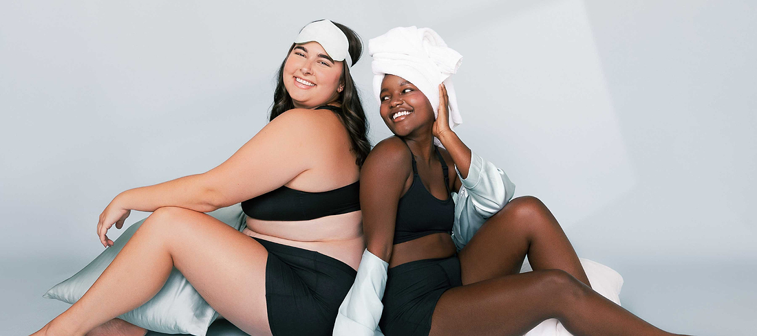 Two women in black loungewear smiling together, one in a sleep mask and the other with a towel wrap.
