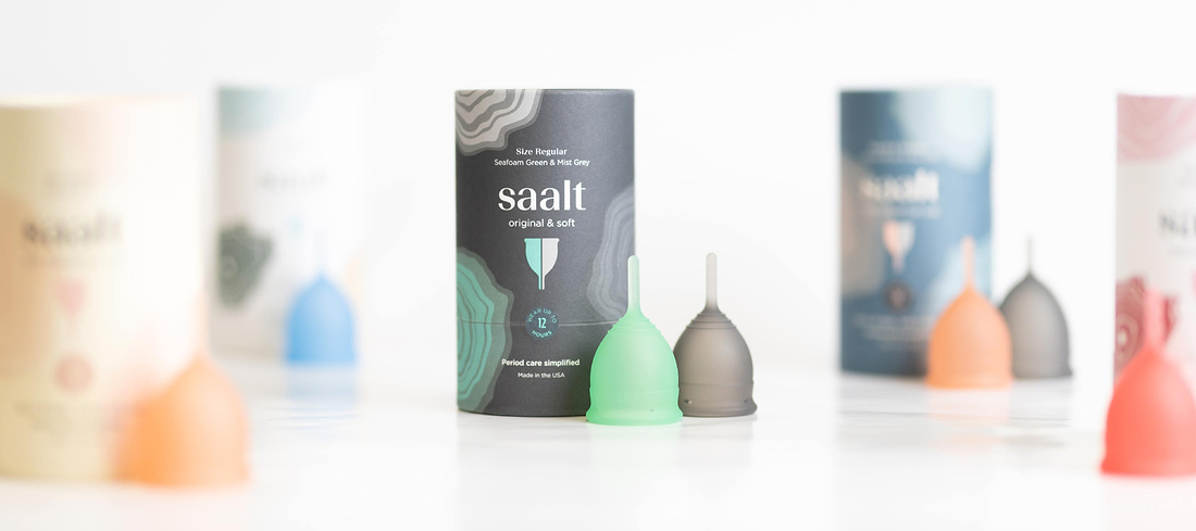 Colorful reusable menstrual cups and packaging displayed on a white surface with blurred products in background.