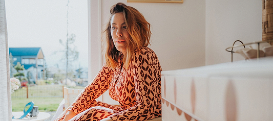 Woman in a leopard-print dress sitting indoors near a sunlit window with a suburban view outside