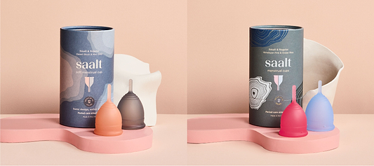 Saalt menstrual cups in pastel colors displayed beside packaging and ceramic bowls on pink platforms.