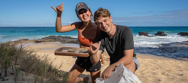 Two people smiling and posing on a sunny beach, holding a tray and bag near the ocean shoreline.