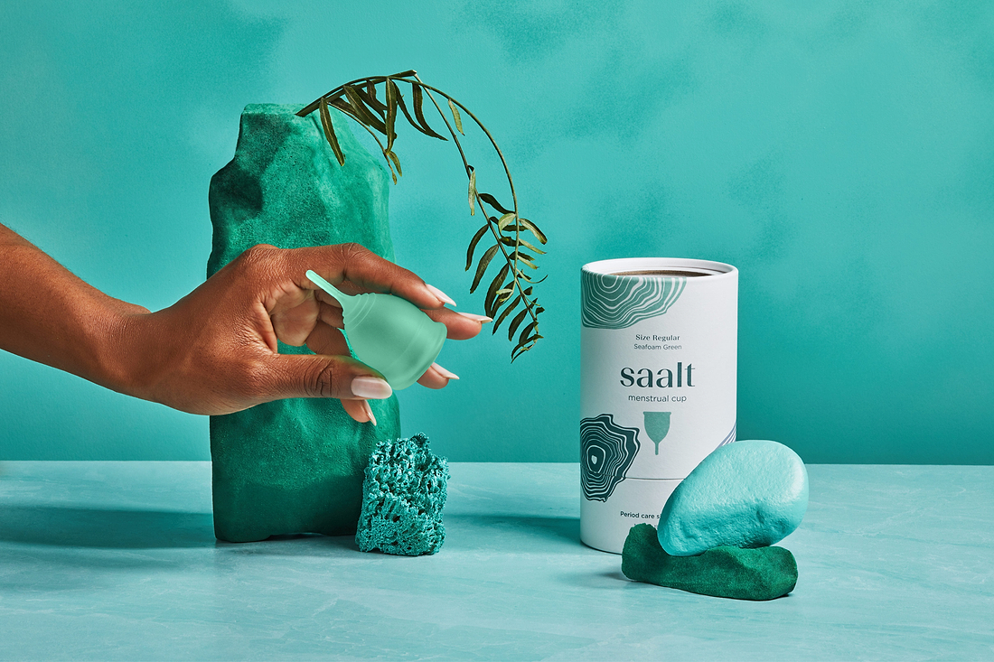 Hand holding a green menstrual cup beside Saalt packaging and decorative teal props.