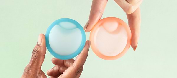 Hands holding two reusable menstrual discs against a soft pastel background.