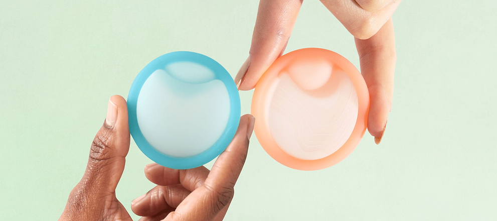 Hands holding two reusable menstrual discs against a soft pastel background.