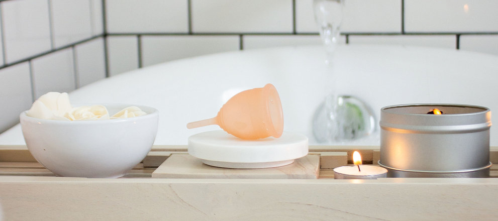 Menstrual cup displayed on a bath tray beside candles and a bowl of cloths in a bathroom.