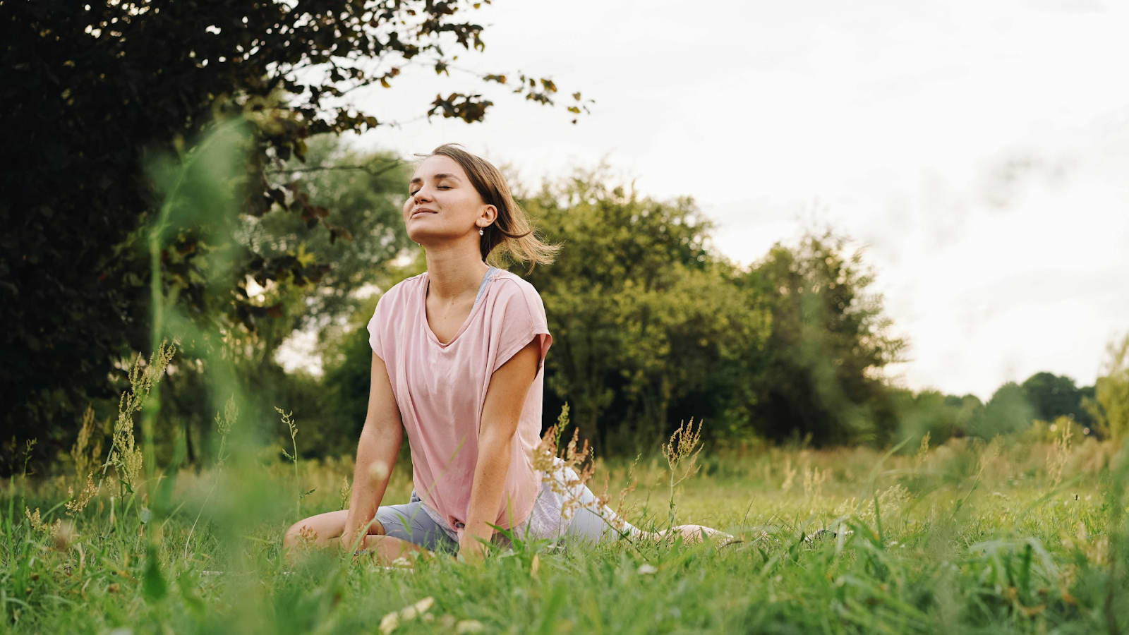 Person sitting peacefully in grassy field outdoors with eyes closed, enjoying nature and fresh air.