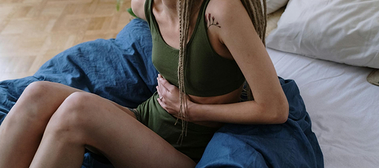 A woman sits on a bed holding her lower abdomen, appearing to experience menstrual cramps or stomach pain.