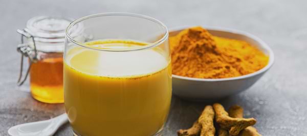 Glass of turmeric milk beside turmeric powder, fresh roots, and a jar of honey on a table.