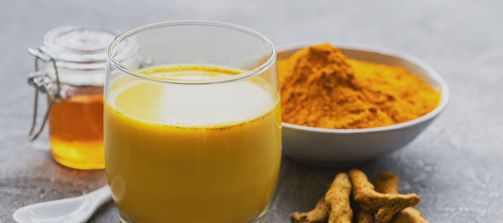 Glass of turmeric milk beside turmeric powder, fresh roots, and a jar of honey on a table.