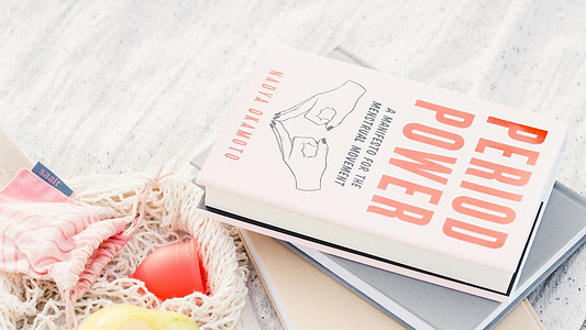 Book titled “Period Power” beside reusable menstrual products and a mesh bag on a light surface.