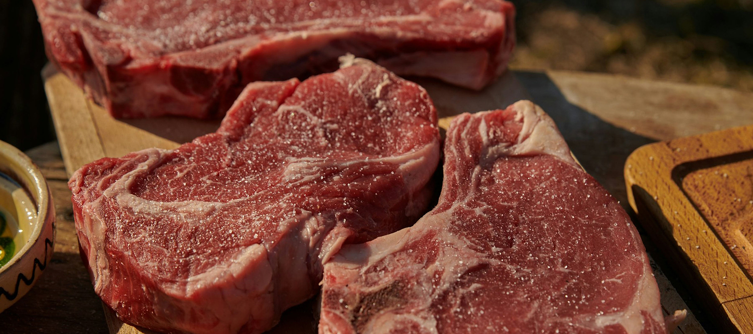Raw bone-in steaks seasoned with salt are displayed on a wooden board outdoors.