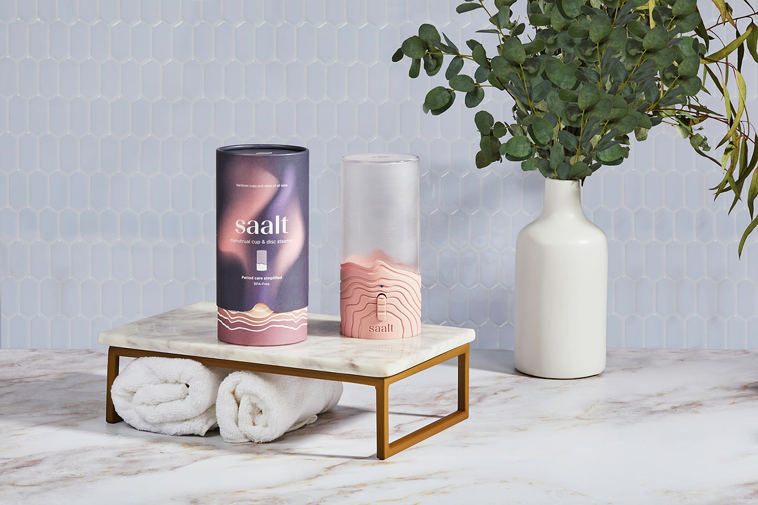 Saalt menstrual cup steamer and packaging displayed on a marble stand beside towels and a vase of greenery