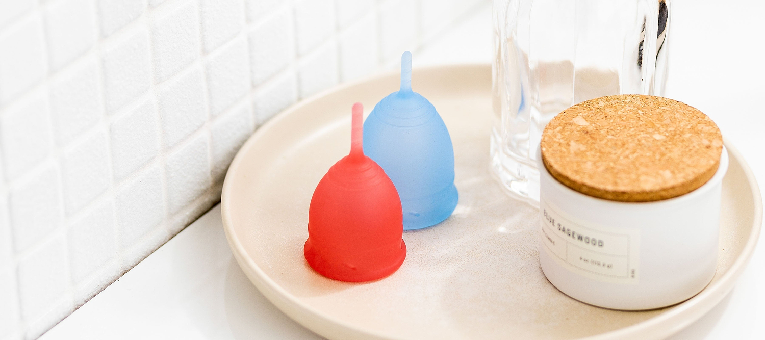 Red and blue menstrual cups on a bathroom tray beside a glass container and candle jar.