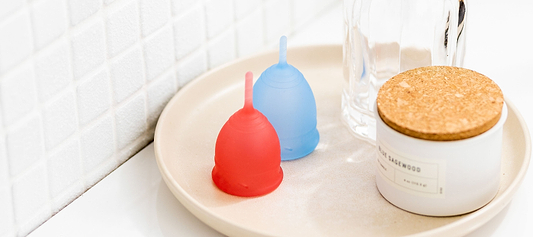 Red and blue menstrual cups on a bathroom tray beside a glass container and candle jar.