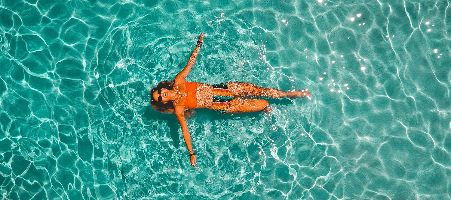 A woman floats relaxed in clear turquoise water, wearing sunglasses and a swimsuit on a sunny day.