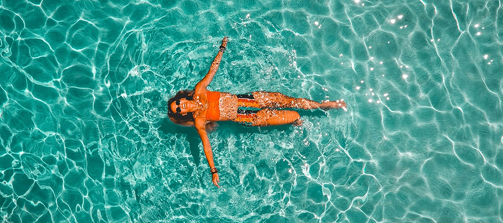 A woman floats relaxed in clear turquoise water, wearing sunglasses and a swimsuit on a sunny day.