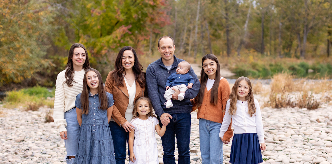 Family of seven posing outdoors on a rocky riverbank with autumn trees in the background