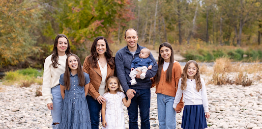 Family of seven posing outdoors on a rocky riverbank with autumn trees in the background