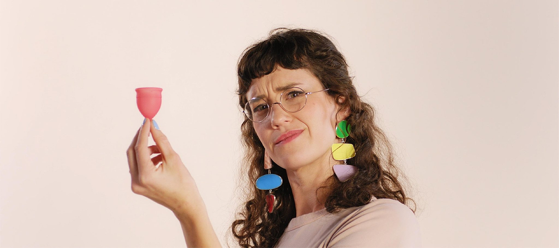 Person wearing colorful earrings and glasses holds a pink menstrual cup with a curious expression