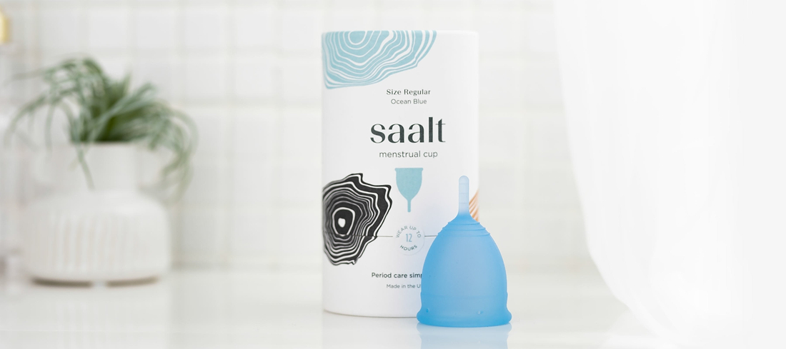 Blue menstrual cup beside Saalt packaging on a bathroom counter with a blurred plant in background.