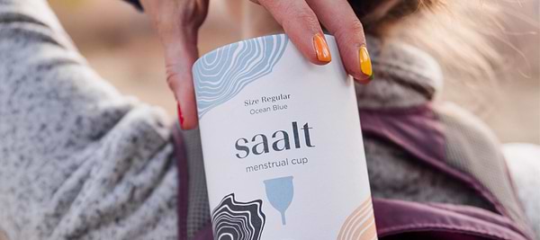 Person holding a Saalt menstrual cup package with colorful painted nails outdoors.
