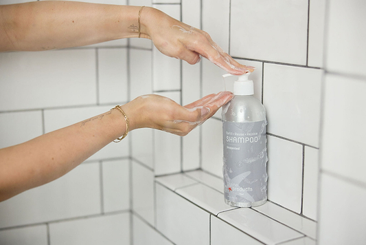 Person dispensing shampoo from a pump bottle in a white tiled shower with soapy hands.