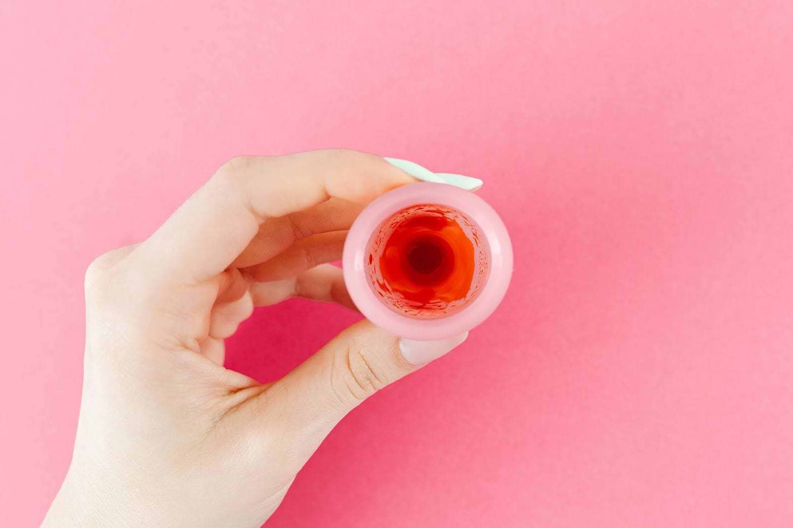 Hand holding a pink menstrual cup against a pink background, viewed from above.