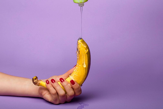 Hand holding a banana with liquid dripping onto it against a purple background, suggestive studio-style image.