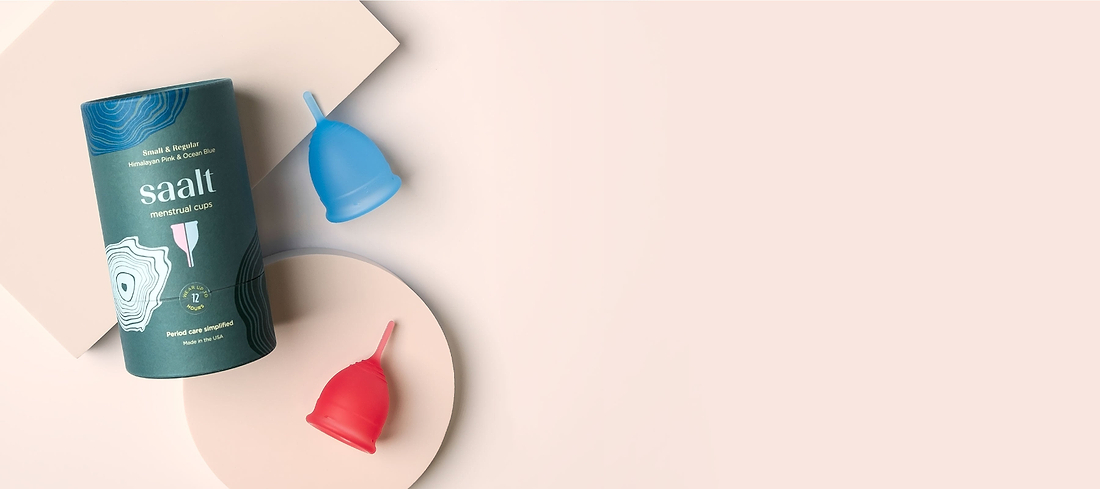 Flat lay of Saalt menstrual cups in pink and blue beside product packaging on a soft beige background.