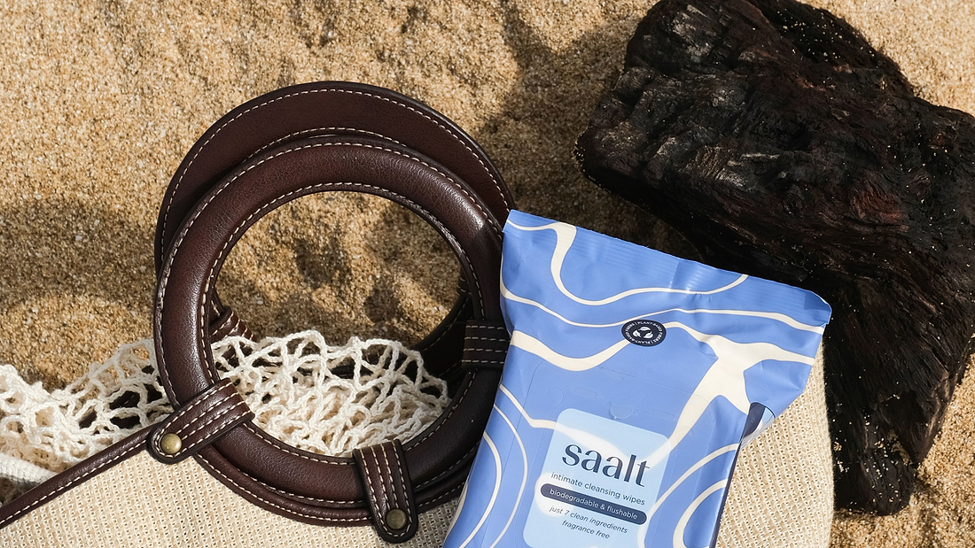 Beach bag with leather handles beside biodegradable intimate cleansing wipes on sandy shore.